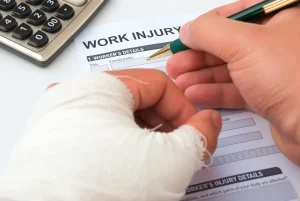 A person with a bandaged hand fills out a work injury report form with a pen, next to a calculator on a desk.