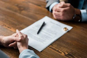 Two people sit across a wooden table from each other with a document, pen, and wedding ring placed between them, suggesting a discussion or negotiation.