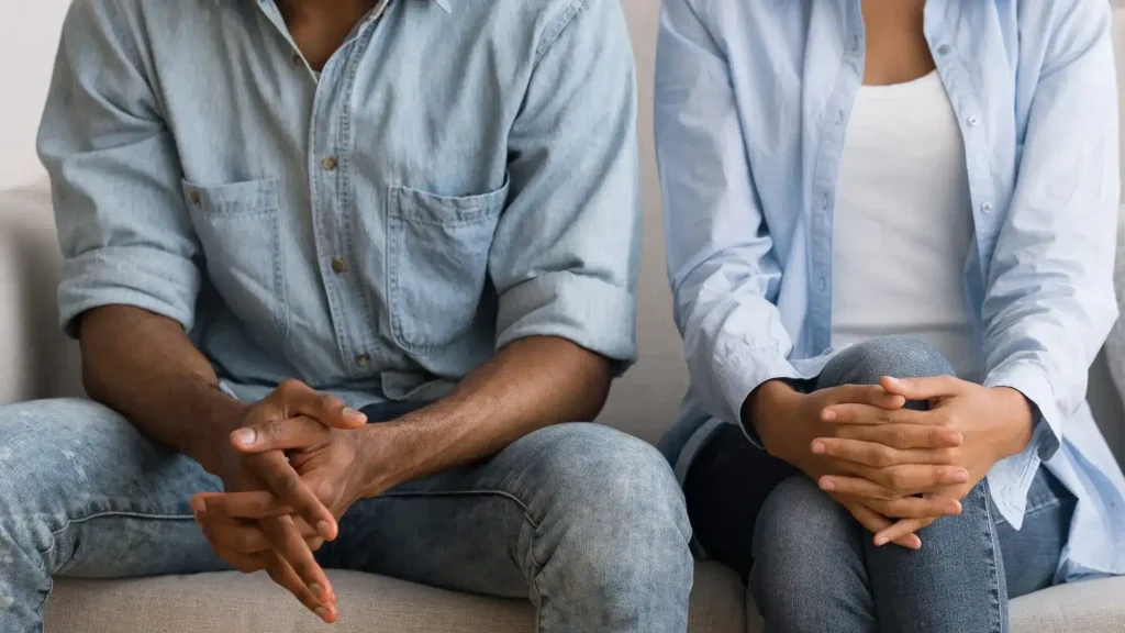 Two people sit side by side on a couch with their hands clasped, both wearing casual blue shirts and jeans. Their faces are not visible.