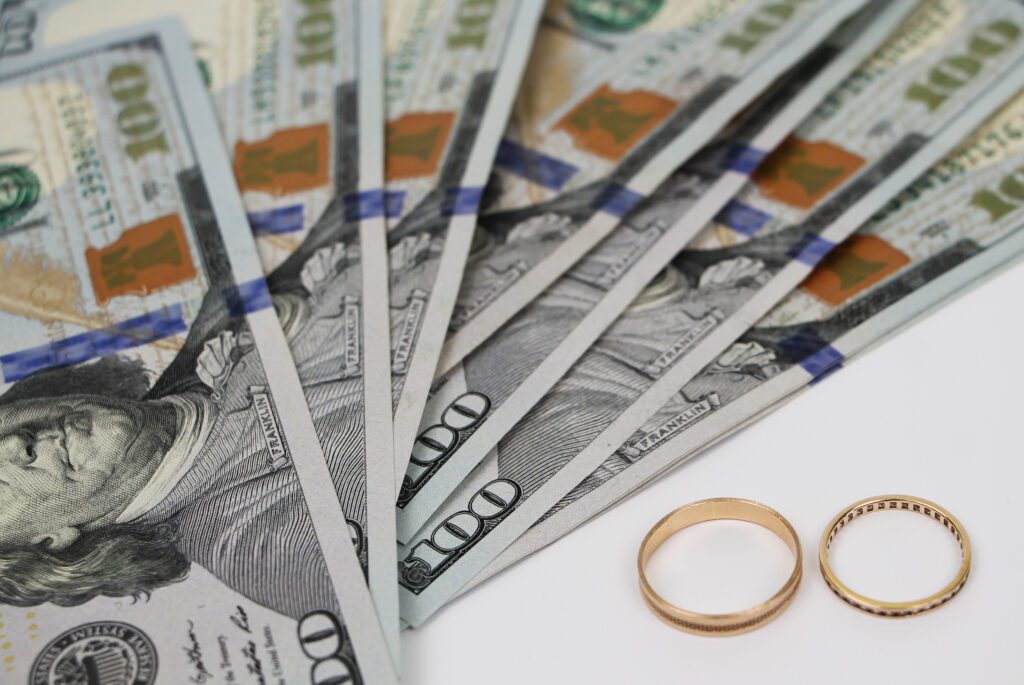Several hundred-dollar bills fanned out next to two gold rings on a white surface.