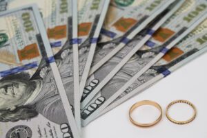 Several hundred-dollar bills fanned out next to two gold rings on a white surface, capturing a moment that might catch the eye of a meticulous Gastonia Divorce Lawyer.
