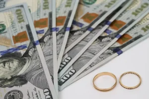 Several $100 bills fanned out next to two gold wedding rings on a white surface.
