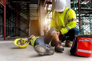 A construction worker in safety gear tends to a colleague lying on the ground with an injured leg. A hard hat and first aid bag are nearby.