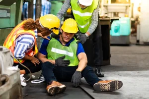 A worker in safety gear sits on the floor holding his stomach while two colleagues assist him in an industrial setting.