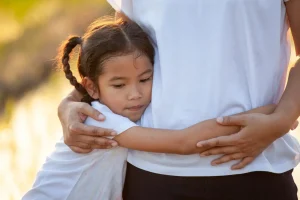 A young girl with braided hair hugs an adult around the waist outdoors, both wearing white shirts. The girl appears thoughtful or pensive.
