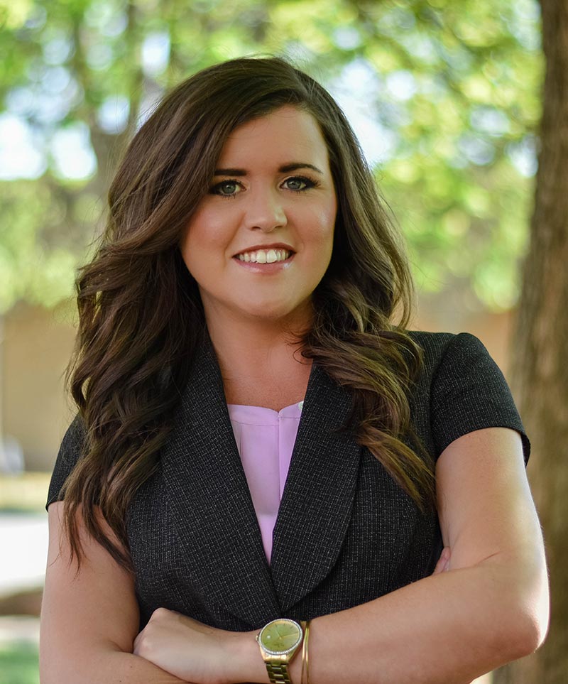 A woman with long brown hair, wearing a dark blazer and light pink top, stands outdoors with her arms crossed, smiling at the camera. Trees in the background create a blurred, leafy backdrop. She exudes confidence and professionalism as a Charlotte Divorce Lawyer.