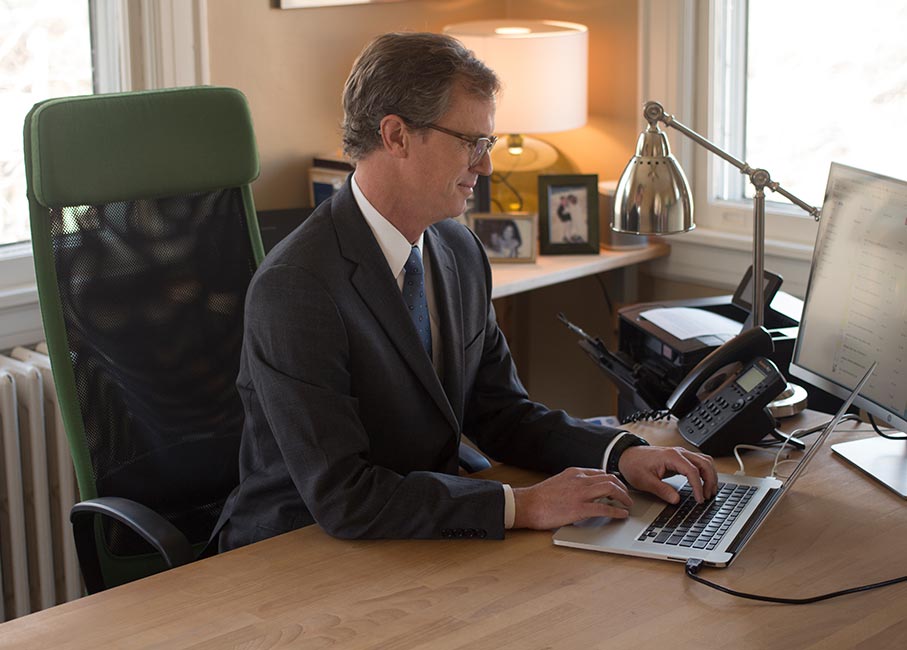A man in a suit sits at a wooden desk, typing on a laptop. The desk, belonging to a Charlotte workers' comp Lawyer, has a telephone, lamp, and framed photos. A window is in the background.