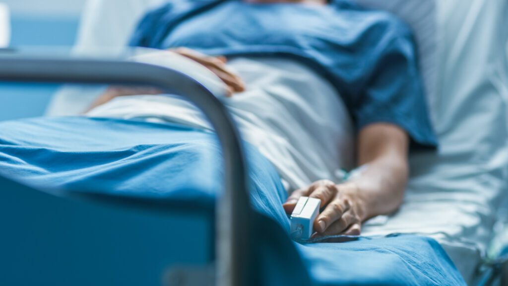 A person lies in a hospital bed, covered by a blue blanket, with a pulse oximeter clipped to their finger. The person's upper body is partially visible.