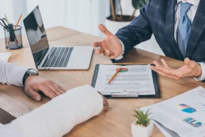 Two people at a desk discuss documents and charts related to Work Comp Benefit; one has a bandaged arm, while the other gestures animatedly. A laptop, clipboard, and papers are spread across the table.