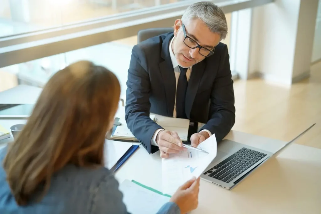 A man in a suit explains Workers' Comp documents to a woman at a desk with a laptop, clipboard, and papers in a modern office setting.