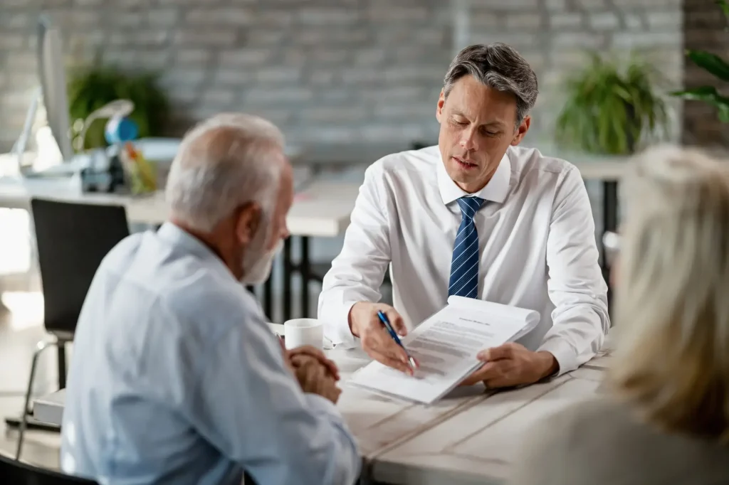 A man in a suit explains a Work Comp Claim document to an older man across a table in a modern office setting, with another person partially visible.