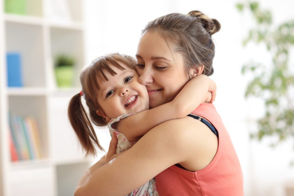 A woman with tied-back hair hugs a smiling young girl with pigtails in a well-lit room, where a bookshelf and potted plant create a cozy backdrop, perhaps highlighting the warmth found in moments of family unity—something valued by any Huntersville Family Law Attorney.