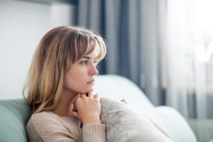 A woman with blonde hair sits on a couch, gazing thoughtfully out a window while holding a cushion close to her chest, contemplating her recent meeting with her Charlotte Workers Compensation Lawyer.