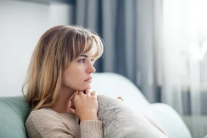 A woman with blonde hair sits on a sofa, resting her chin on her hands and looking thoughtfully out of a window, reflecting on her experience with domestic violence.