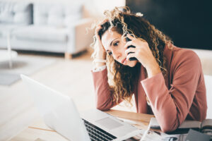 Cyrly busy business woman suffering stress working at laptop while talking on mobile phone in overwork. Tired businesswoman using phone. Girl feeling sad.
