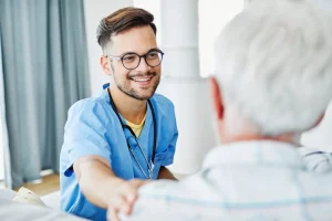 A young male nurse in blue scrubs smiles and supports an older male patient, placing a hand on his shoulder during a conversation.