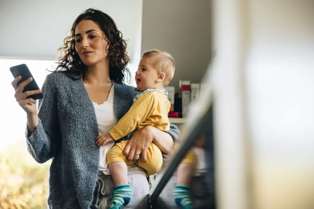 A woman holding a baby in one arm looks at her smartphone in the other, managing parallel parenting tasks while standing indoors.