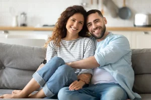 A smiling couple sits closely together on a sofa in a modern kitchen, both wearing casual clothes and looking at the camera, showcasing harmony even after divorce.
