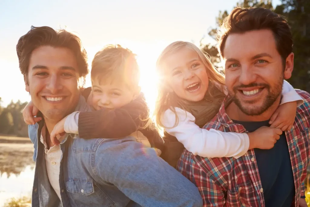 Two same-sex men outdoors give piggyback rides to a young boy and girl, all smiling at the camera with sunlight in the background.