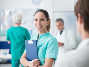 A nurse in teal scrubs holds a clipboard and smiles at the camera in a medical office, where healthcare professionals support patients with Workers' Compensation needs.