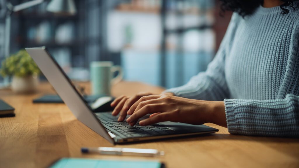 A person wearing a blue sweater types on a laptop at a wooden desk with a pen, notepad, and coffee mug nearby, perhaps researching the best Charlotte Workers Compensation Lawyer.