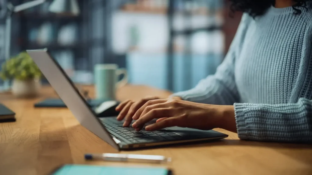 An independent contractor in a blue sweater types on a laptop at a wooden desk, with a mug and office supplies visible in the background.
