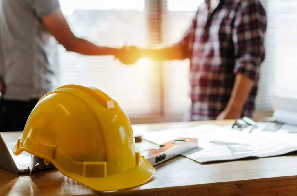 Construction hard hat on a desk with workers shaking hands in the background during a workers’ compensation or workplace injury discussion.