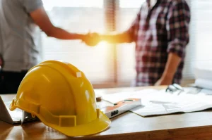 A yellow safety helmet rests on a desk with papers and tools, capturing the essence of a construction site as two people shake hands in the background.