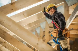 Construction worker wearing safety gear stands on wooden rafters, reaching upward with one hand while holding a hammer in the other. Sunlight illuminates the partially built roof structure—highlighting risks often present in a fatal workplace.