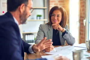 Two professionally dressed people sit at a desk having a conversation about a settlement, with documents and a pen on the table in front of them.