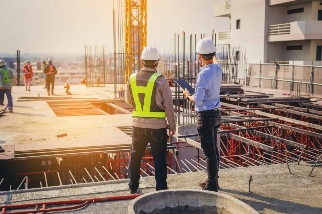 Two construction workers in safety gear stand on a building site, discussing plans while others work in the background amid scaffolding and building materials, staying alert to prevent Charlotte construction work accidents.