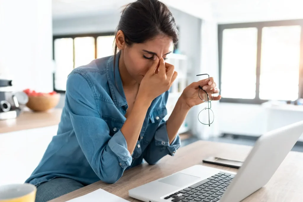 A woman sitting at a desk in front of a laptop, holding her glasses and rubbing her eyes, appears stressed or tired—possibly due to the pressures of dealing with divorce.