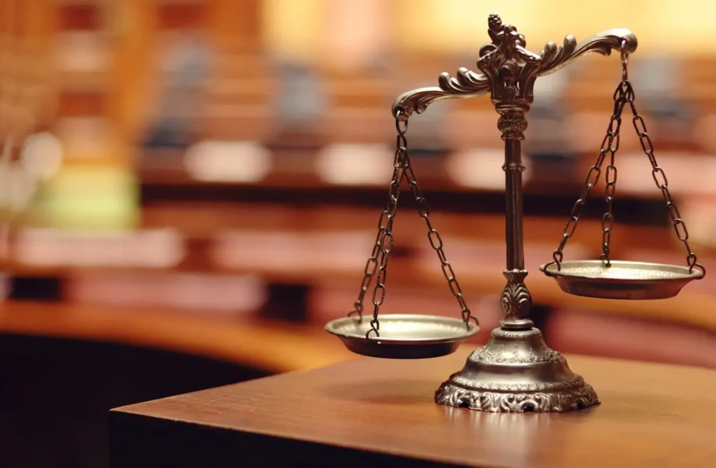 A close-up of a bronze balance scale on a wooden desk in a courtroom, with blurred rows of seats and warm lighting in the background, evokes the careful deliberation seen in every workers comp settlement chart.