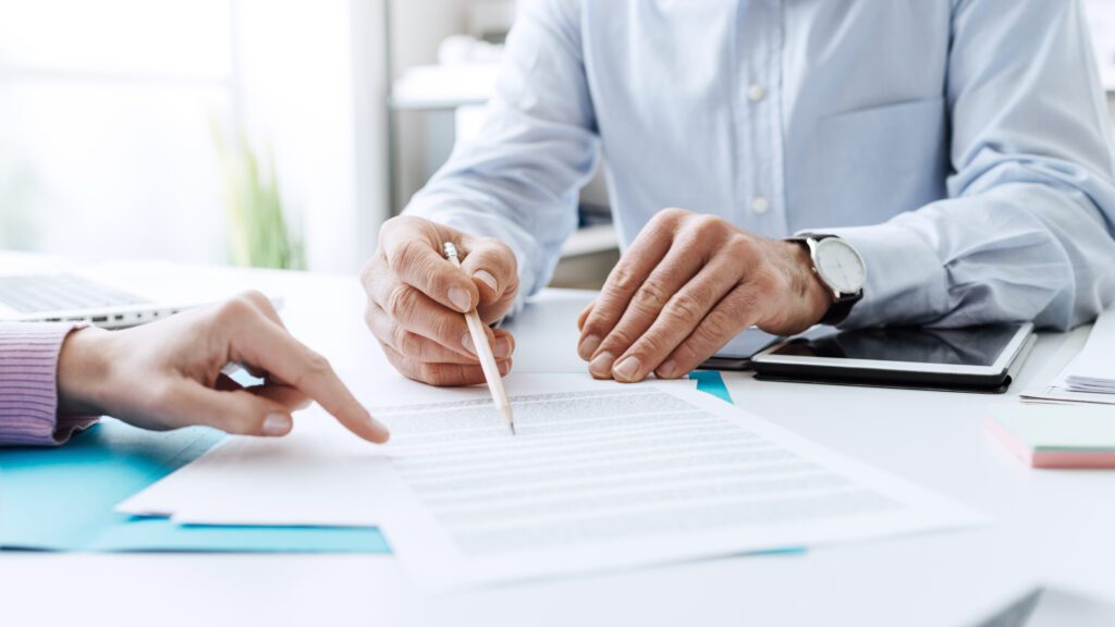 Two people are reviewing a document on a desk cluttered with papers and a tablet. One person points to the document, while the other holds a pencil, indicating collaboration and discussion—much like what you might see with Charlotte high asset divorce Lawyers working together on a complex case.