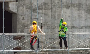 Two construction workers wearing safety gear and helmets work on scaffolding in front of a concrete wall at a North Carolina job site.