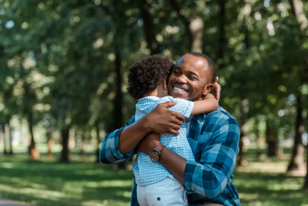 A man wearing a blue plaid shirt hugs a young child outdoors in a park with trees in the background, capturing a heartfelt family moment that a Charlotte father's rights lawyer helps protect.