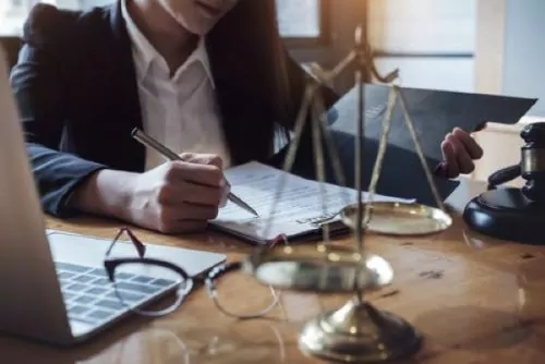 A person in business attire is writing in a notepad at a desk, surrounded by a laptop, scales of justice, glasses, and a gavel. The scene could easily depict the office of a seasoned Charlotte delivery driver Workers Compensation Lawyer diligently preparing for an important case.