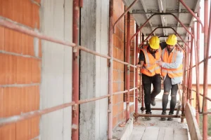 Two construction workers in safety vests and helmets walk on scaffolding; one appears to assist the other, who is suffering a workplace injury and limping.