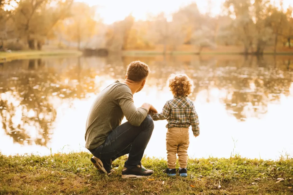 An adult and a child stand on grass by a lake, facing the water, with trees and soft sunlight in the background—capturing a peaceful moment that might comfort those seeking guidance from a child custody lawyer in Charlotte.