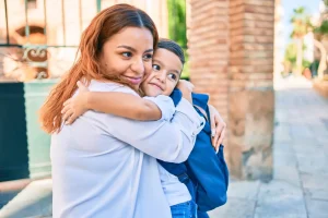 A woman hugs a smiling young boy with a backpack outdoors near a brick wall on a sunny day, capturing the warmth and support that family lawyers strive to protect every day.