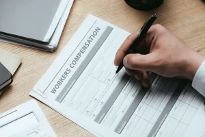 A person fills out a Charlotte workers comp claim form on a desk with a pen, next to documents and a calculator.