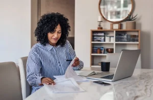 Person sitting at a table with a laptop, reviewing Charlotte workers compensation papers and holding a pen, with a coffee mug, smartphone, and shelves in the background.
