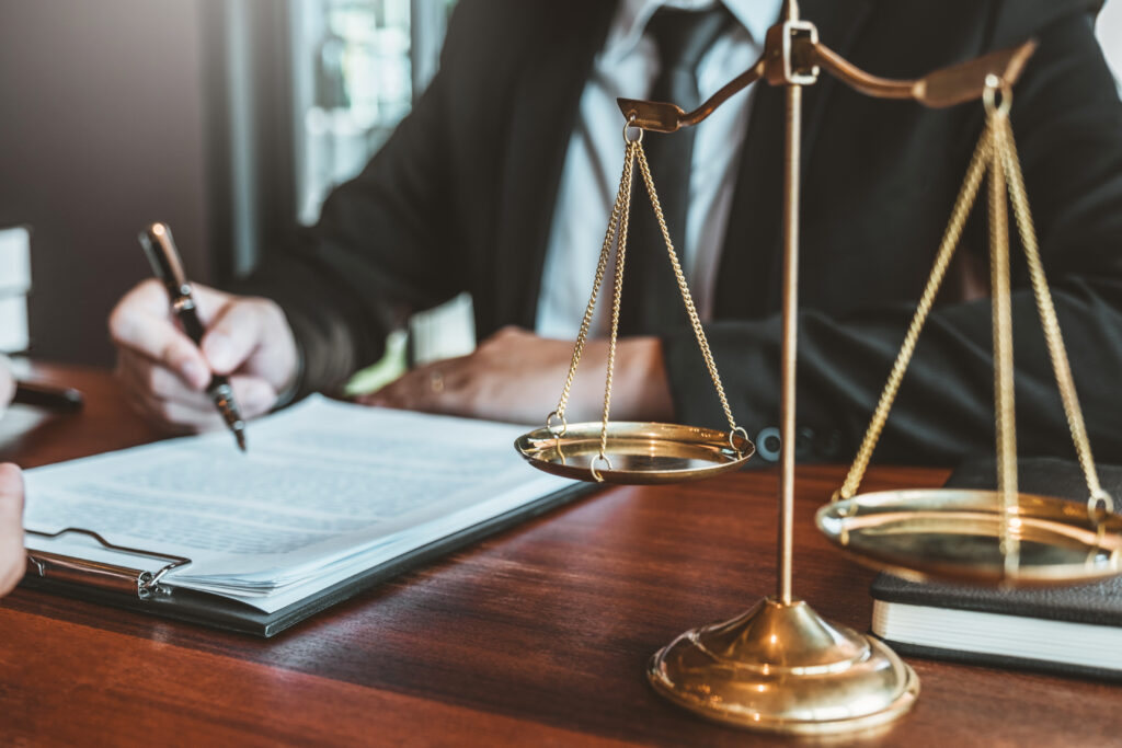 A lawyer is seen signing a document at a desk, with a golden balance scale prominently displayed in the foreground.