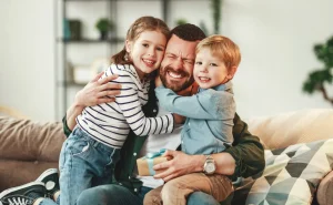 A man sitting on a couch hugs two smiling children while holding a small gift box in a living room, sharing a joyful moment that highlights the importance of family—something every Charlotte family law attorney understands deeply.