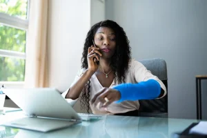 A woman with a blue arm cast sits at a desk, talking on the phone and using a laptop, possibly handling workers comp claims.