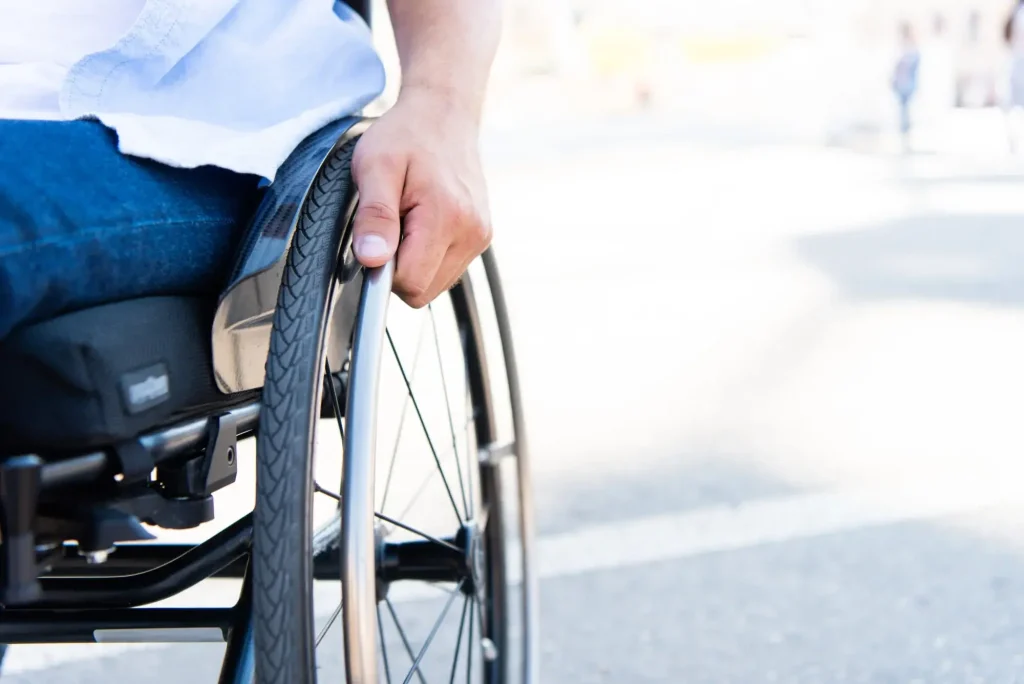 A person wearing blue jeans and a white shirt propels a manual wheelchair outdoors, highlighting their hand on the wheel—an image often associated with workers comp and disability support.