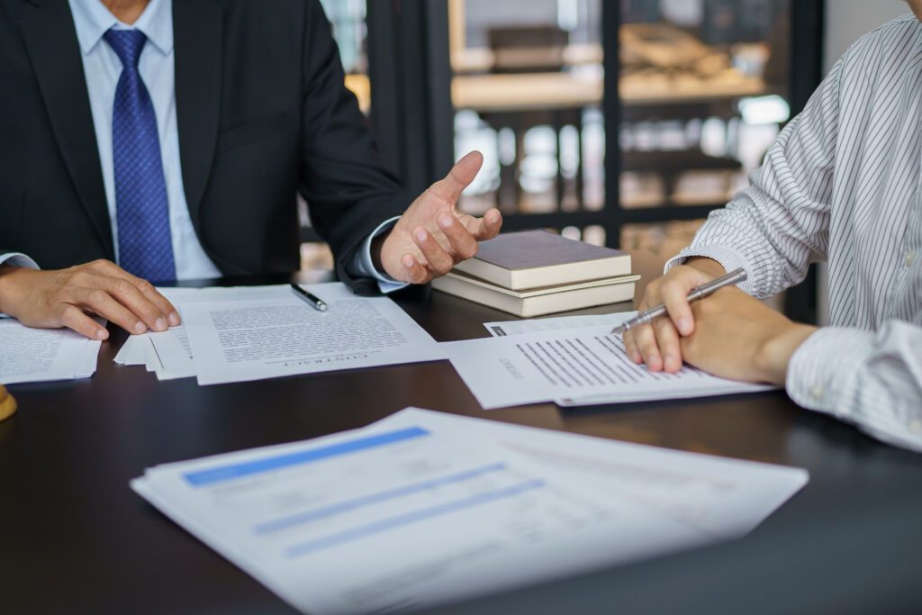 Two individuals are seated at a table with documents, notebooks, and a pen. One person gestures with their hand while the other writes on a piece of paper. It appears they are discussing legal matters.