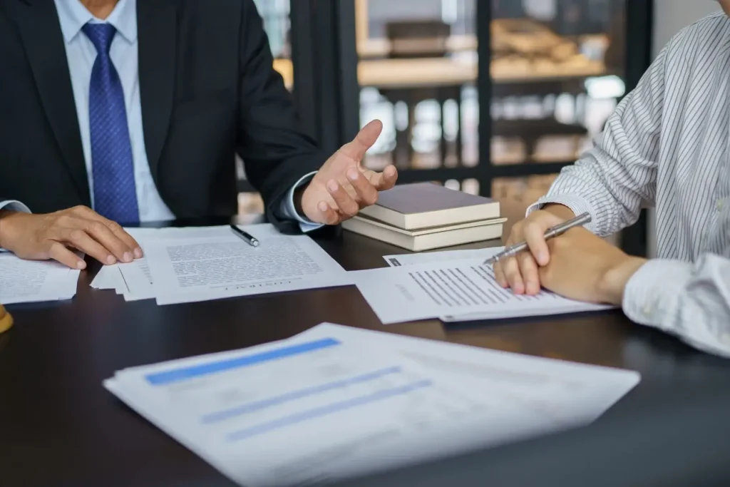 Two people sit at a desk in an office, discussing documents related to workers comp in North Carolina; one wears a business suit, while the other writes notes on a clipboard.