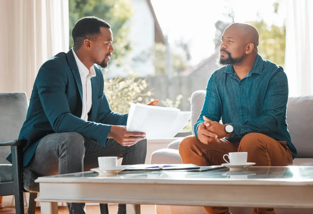 Two men sit on a sofa in a well-lit room, discussing important matters. One holds papers and gestures while the other listens intently. Two cups and important documents are on the table in front of them.