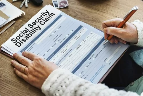 A person is meticulously filling out a Social Security Disability Claim form on a wooden desk, surrounded by keys, a photo, and other relevant documents.